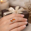 Female hand wearing a sterling silver bypass ring with two oval honey Baltic amber stones, photographed in a warm lifestyle setting with soft fabric, candle, and gift box in the background.