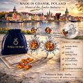Promotional image for Holiday Gift Shops featuring sterling silver sun-shaped amber drop earrings against a scenic waterfront view of Gdańsk, Poland at sunset. Text highlights “Made in Gdańsk, Poland — Heart of the Amber Industry,” with product callouts showing post back closure, genuine Baltic amber, solid 925 sterling silver, lightweight comfort, approximate weight of 3.6 g, and certificate included. A blue Holiday Gift Shops pouch, certificate of authenticity, and loose amber stones are displayed in the foreground.