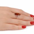 Hand wearing an oval cognac amber ring in a slim sterling silver setting, shown on a finger with glossy red nail polish against a light background.