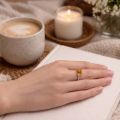 A close-up lifestyle photo of a hand resting on a light fabric-covered book, wearing a silver ring with a square golden-yellow gemstone. In the softly blurred background are a cup of coffee, a lit candle on a woven coaster, and small white flowers, creating a warm, cozy atmosphere.